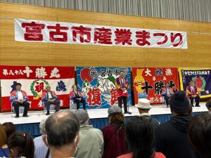 「宮古市産業まつり」と書かれた横断幕が吊るされ、複数の大漁旗が壁に張られた舞台状で、法被を着た参加者たちが横一列に椅子に座って並び、中央の男性がスタンドマイクの前で話をしている様子の写真