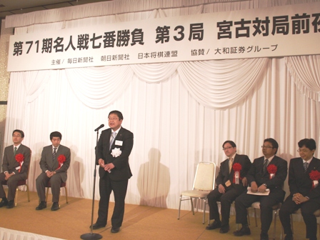 「第71期名人戦七番勝負 第3局 宮古対局前夜祭」の吊り看板があり、ステージの壁際に座る関係者とマイクスタンドの前に立つ市長の写真