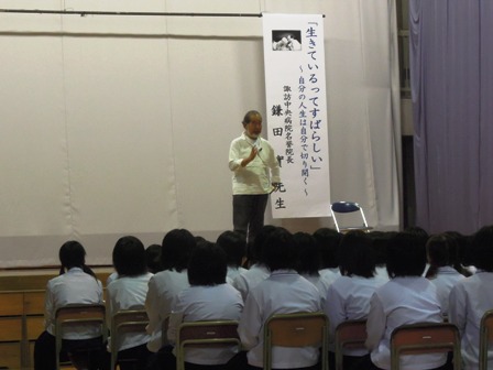 「生きているってすばらしい」と書かれた横断幕が飾られた舞台上で話をする鎌田實先生と、椅子に座り話を聞く生徒達の写真