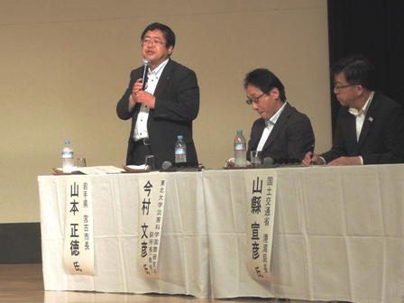 ステージ上に設置された白い布をかけた長机の前に男性2名が座り、市長は立ってマイクを持ち話をしている写真