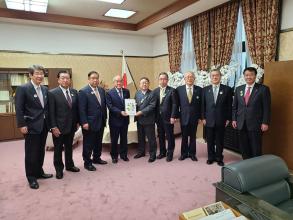 国旗が置かれた室内で、鈴木俊一財務大臣と市長が要望書を一緒に持ち、7名の関係者の方々が横一列に並んでいる写真