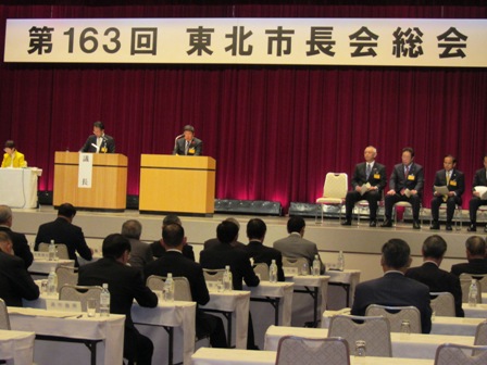 「第163回 東北市長会総会」と書かれた横断幕が吊るされた舞台上で、椅子に座る関係者や2台置かれた演台の前に立つ男性2名と、会場に置かれた長机の席に着く関係者の方々の写真