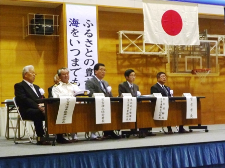 「ふるさとの豊かな海をいつまでも」と書かれた垂れ幕と日の丸の前に5名の男性が舞台上に置かれた長机に座っている写真