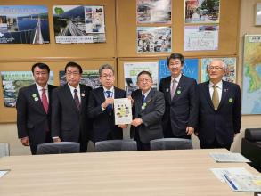 ポスターが貼られた掲示板の前で、国土交通省の村山一弥道路局長と市長が要望書を一緒に持ち、4名の関係者の方々が横一列に並んでいる写真