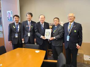 黒色の椅子が設置されたテーブルの傍で、国会議員の佐藤信秋参議院議員と市長が要望書を一緒に持ち、3名の関係者の方々が横一列に並んでいる写真