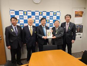 壁にバックパネルが設置された室内で、国会議員の足立敏之参議院議員と市長が要望書を一緒に持ち、3名の関係者の方々が横一列に並んでいる写真