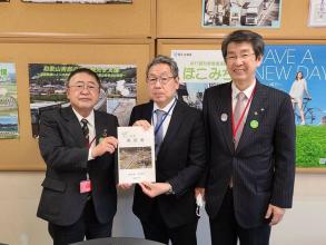 ポスターが貼られた掲示板の前で、国土交通省の村山一弥道路局長と市長が一緒に要望書を持ち、山田町の佐藤信逸町長と横一列に並んでいる写真