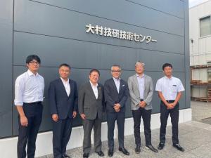 黒を基調とした外壁の「大村技研技術センター」と書かれた切文字看板の前に市長や大村技研株式会社の関係者ら6名が並んでいる写真