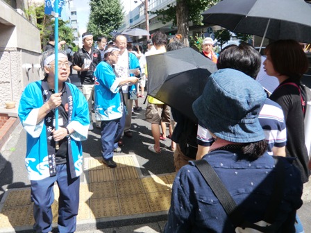 並んでいるお客様の横を水色に渦潮模様の半被をきた男性がのぼり旗を持ちながら歩いている写真