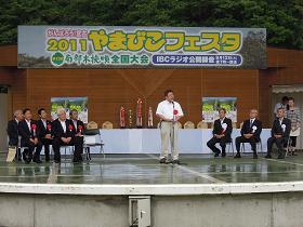 「2011やまびこフェスタ」と書かれた吊り看板のステージ上に、来賓者が両側に座っており、中央のマイクスタンドの前に立つ市長が挨拶を行っている様子の写真