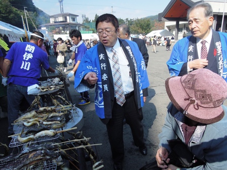大空の下、新里まつりで賑わっている中、青いはっぴを着た市長と関係者が、網の上で串焼きの魚に向けて指をさしている写真