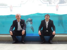品川水族館のイルカショーの水槽前に座る市長と男性の間に、水槽越しにイルカが映っている写真