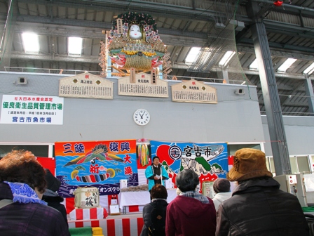 日本一の大熊手が飾られた宮古市魚市場内に大漁旗が飾られ、水色のはっぴを着た市長がマイクスタンドの前に立っている写真