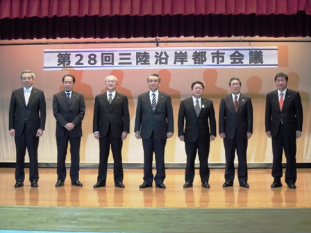 「第28回三陸沿岸都市会議」と書かれた舞台看板があるステージ上にスーツを着た男性7名が立ってる写真