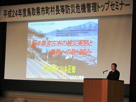 「平成24年度鳥取県市町村長等防災危機管理トップセミナー」の吊り看板があり、「岩手県宮古市の災害実態と復興への取り組み」と題した資料がスクリーンに投影されており、演壇で市長が講演している写真