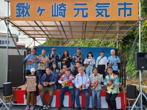 「鍬ヶ崎元気市」と書かれた小さなステージに、市長と副市長を含む5名が座り、後列に8名のウクレレを持った参加者が並んでいる写真