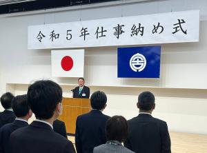 令和5年仕事納め式とかかれた幕が飾られ、並んでいる職員にむかって演台から挨拶をしている市長の写真