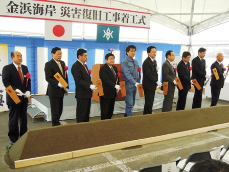 「金浜海岸災害復旧工事着工式」と書かれた横断幕の前でスーツと作業着をきた男性達が鍬を持ち並んでいる写真