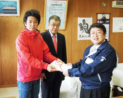 赤い服にジーンズをはいた男性と市長が金封を一緒に持ち記念撮影している写真
