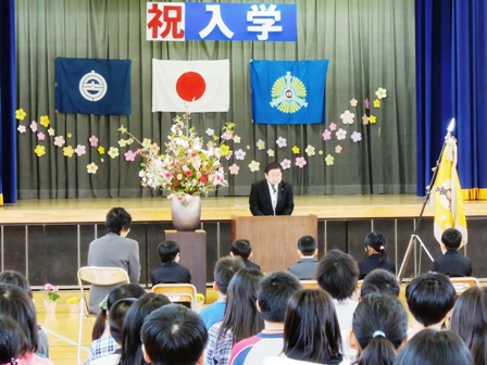 舞台に「祝入学」と書かれた舞台看板があり、舞台下には、大きな花が飾られその横の演壇で市長が座っている新入生に話をしている写真