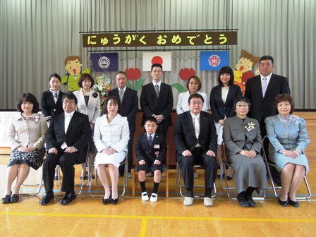 「にゅうがくおめでとう」と掲げられた吊り看板の舞台前で、市長と入学した一年生、関係者達が横2列に並んでいる写真