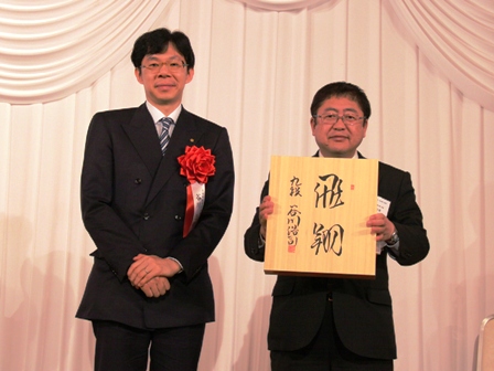 「飛翔」と書かれた将棋盤を持つ市長と、谷川浩司会長が横に並んでいる写真