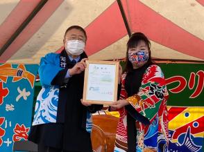 鮭・あわびまつり」会場のステージ上で、水色の法被を着た市長と柄入りの法被を着た演歌歌手の三本木智子さんが額に入った賞状を一緒に持っている写真