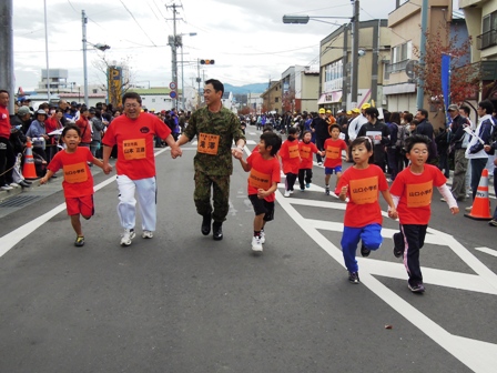 赤いTシャツを着た市長と迷彩柄の服を着た滝澤博文第2師団幕僚長がペアの子供たちと手をつなぎ道路を走っており、沿道からは多くの観客が声援を送っている写真