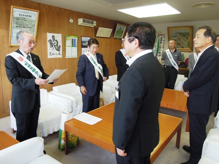 市長の向かい側にたすきをかけ賞状を読み上げているスーツ姿の男性と周りに関係者が立ち並んでいる室内の写真