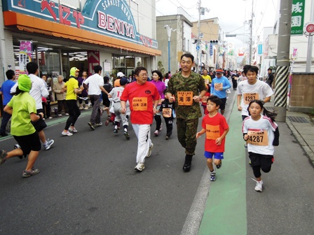 商店街を市長、滝澤幕僚長、子供達が走り折り返し地点から戻ってきた方とすれ違っている写真