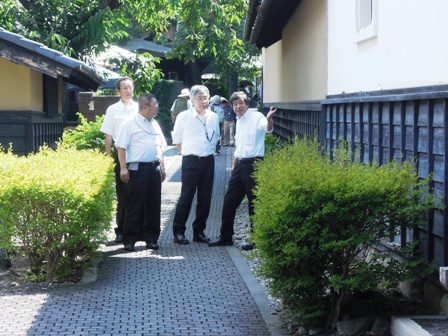 建物の下半分は正方形の黒い板が敷き詰められ、上半分が白く塗られた建物と栗の木を敷き詰めた歩道でスーツを着た男性が3名の男性に建物の説明をしている写真