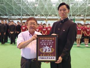 市長と学ランを着た男性が額に入れられた記念品のようなものを持ち、その後ろにたくさんの応援部員が並んでいる写真
