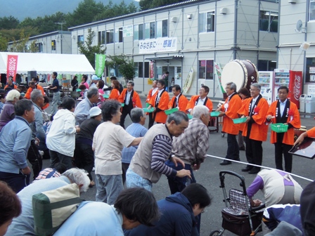 2階建ての仮説店舗が立ち並ぶ入り口前の敷地内で、オレンジのはっぴを着た市長や関係者が餅まきの餅を投げており、市民の皆さんが手を伸ばし賑わっている様子の写真