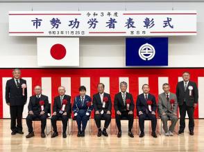 「令和3年度市勢功労者表彰式」と書かれた横断幕と2つの旗、紅白幕が飾られた会場に設置された椅子に胸章リボンを付けた7名の受賞者が横一列に並んで座り、右端に市長と左端に関係者の男性が立っている記念写真