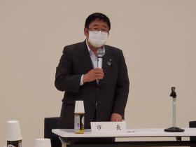 市長の名札が置かれた席で立ち、歯科保健事業の実施内容と事業計画について説明している写真