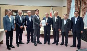 日本国旗の前方でスーツを着た8名の男性が横一列に並び、中央の2名が要望書を一緒に持っている記念写真