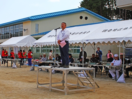 中学校のグランドに白いテントが立ち並び、関係者が参列し、朝礼台でマイクスタンドの前に立つ市長の写真