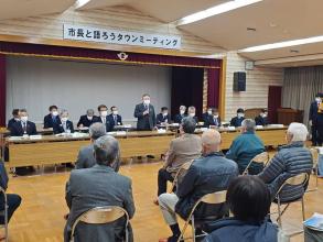 小さなステージの上に「市長と語ろうタウンミーティング」と書かれた横断幕が掲げられた会場で、2列に設置された長机の席に座った関係者とパイプ椅子に座った参加者が向かい合って座り、中央の席から立ち上がった市長が挨拶を行っている川井会場の様子の写真