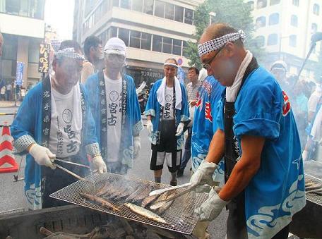 青色の半被に鉢巻をまいた男性達が網の上でさんまを焼いている写真