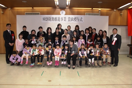 「8020運動推進事業賞状授与式」と書かれた横断幕の下、3～4歳くらいの子供たちが白いリボンを胸につけて、横並びで2列に並び、後方に市長や関係者と保護者が並んでいる写真