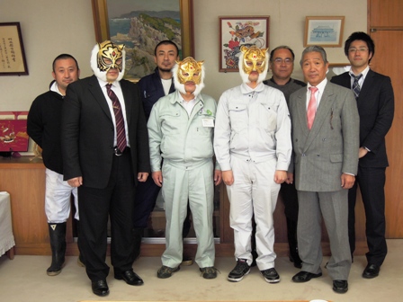 スーツや作業着をきた男性8名が並び、そのうち3人は、プロレスマスクを被り記念撮影している写真