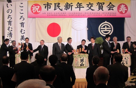 「市民新年交賀会」の吊り看板があり、舞台上に置かれた樽酒を、市長と関係者が木槌を持って鏡開きをしており、参加者が鏡開きの様子を舞台下から見ている写真