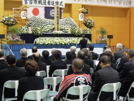 戦没者追悼式の祭壇の前で、追悼の言葉を述べている山本市長とそれを座って聞いている参加者の方々の写真