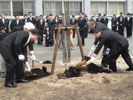 スーツを着た男性達が地面に穴が掘られ木が植えられた根元に土をスコップで入れているのを参加者達が見ている写真