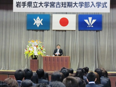 「岩手県立大学宮古短期大学部入学式」と書かれた吊り看板があり、縦に整列する生徒達の前で、演壇に立ち祝辞を述べる市長の写真