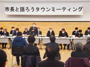 前方の壁沿いに「市長と語ろうタウンミーティング」と書かれた横断幕が掲げられた会場で、2列に設置された長机の席に座った関係者とパイプ椅子に座った参加者が向かい合って座り、中央の席から立ち上がったマイクを持った市長が挨拶を行っている宮古会場の様子の写真