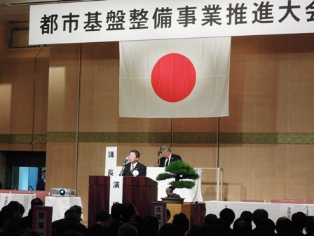 「都市基盤整備事業推進大会」の吊り看板に、日の丸の国旗が飾られており、市長が演壇に立って話をしている写真