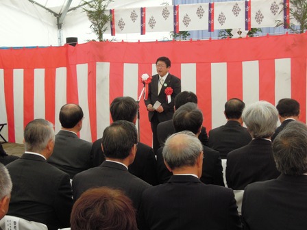 紅白幕が張られた安全祈願祭の会場で、マイクスタンドの前に立つ市長と、椅子に座る関係者の写真