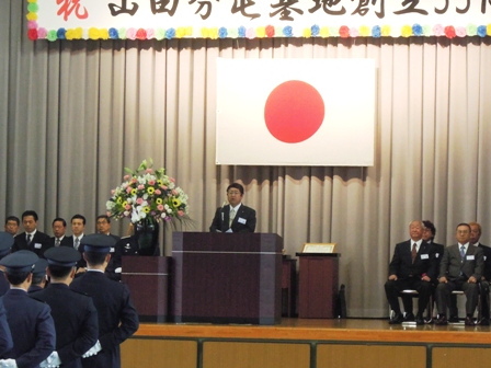 日の丸の旗がかけられたステージ上の演壇でスーツを着た男性と両隣りに並べられたパイプ椅子に複数の男性が座っている写真