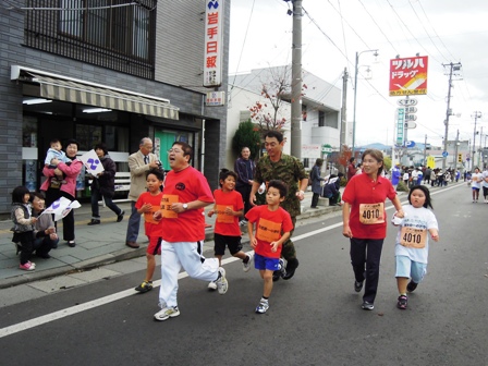 歩道で応援して旗を振ってくれている方々と商店街を市長、子供達、滝澤幕僚長が走っている写真